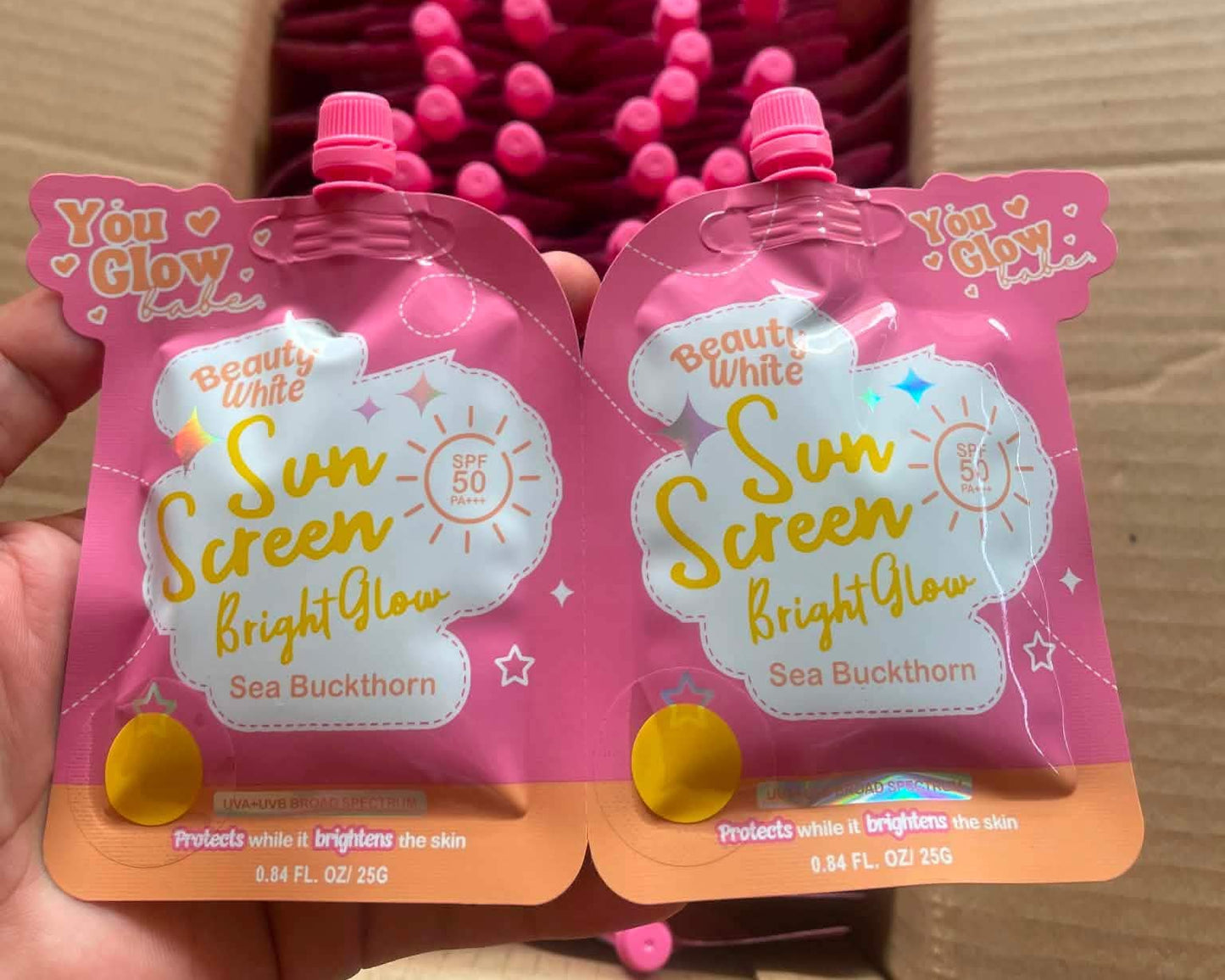 Two pink sun screen pouches with visible branding held in front of a box filled with similar products.