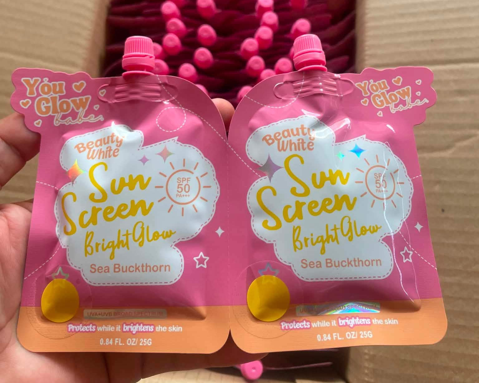 Two pink sun screen pouches with visible branding held in front of a box filled with similar products.
